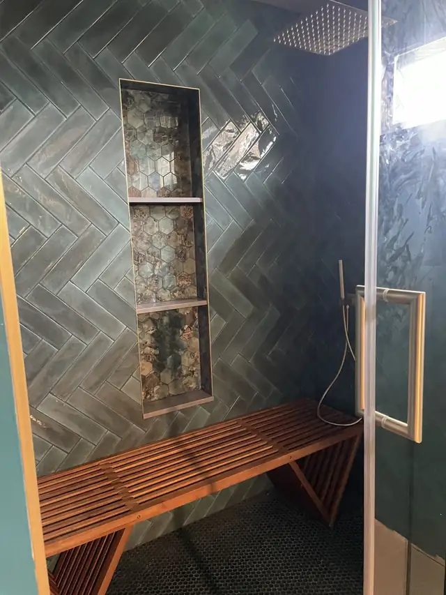 Green herringbone tile shower with niche, wood slat bench and frameless glass door