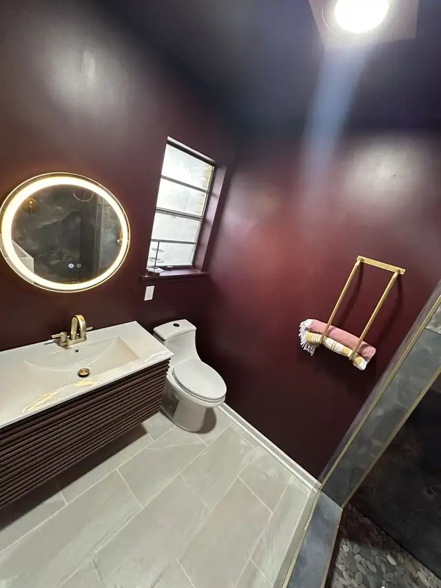Fluted wood floating vanity with a white top, lighted round mirror and brass towel ladder in a burgundy bathroom