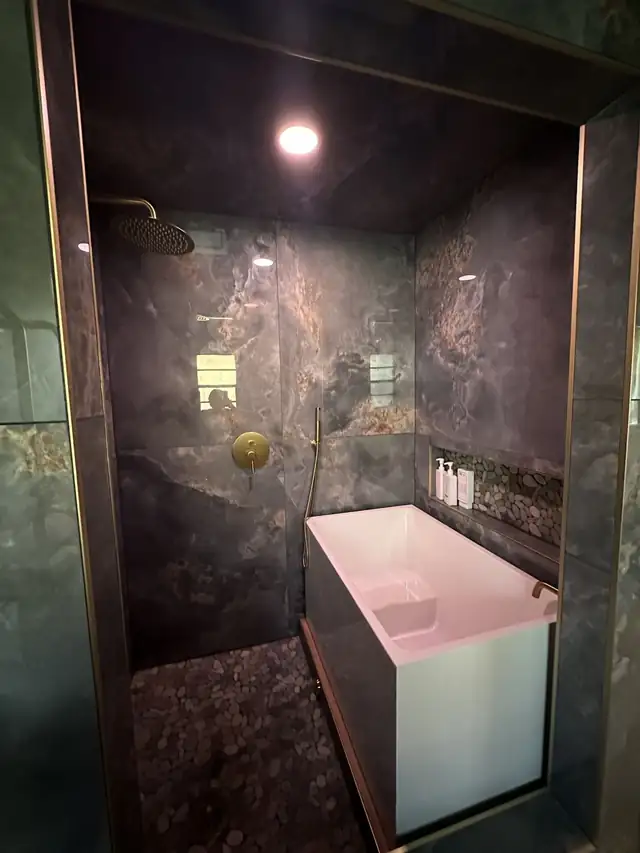Remodeled bathroom with dark stone-look shower walls, a white soaking tub and a brushed brass shower set