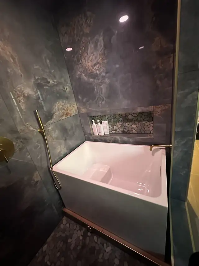 White square soaking tub with a brushed brass shower wand and a stone-look wall niche